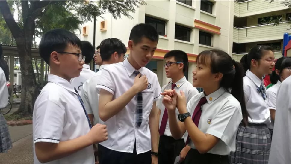 Several school students in white uniforms with ties chat outdoors near a multi-story building.